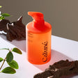 Immagine prodotto del Cafuné Shampoo scalp care: il flacone arancione con erogatore pump è ambientato in un contesto naturale con legni e foglie verdi, richiamando gli attivi botanici della formula. Visual ideale per comunicare detersione delicata, equilibrio del sebo e cura del cuoio capelluto.