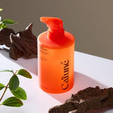 Immagine prodotto del Cafuné Shampoo scalp care: il flacone arancione con erogatore pump è ambientato in un contesto naturale con legni e foglie verdi, richiamando gli attivi botanici della formula. Visual ideale per comunicare detersione delicata, equilibrio del sebo e cura del cuoio capelluto.