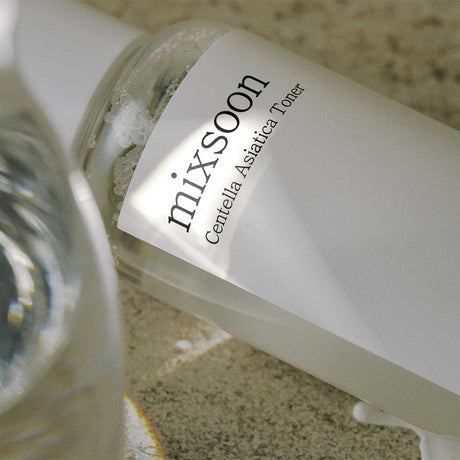 Primo piano della bottiglia Mixsoon Centella Asiatica Toner adagiata su una superficie naturale. La luce morbida e i riflessi nell’acqua enfatizzano la purezza e la trasparenza del tonico calmante per la pelle.

