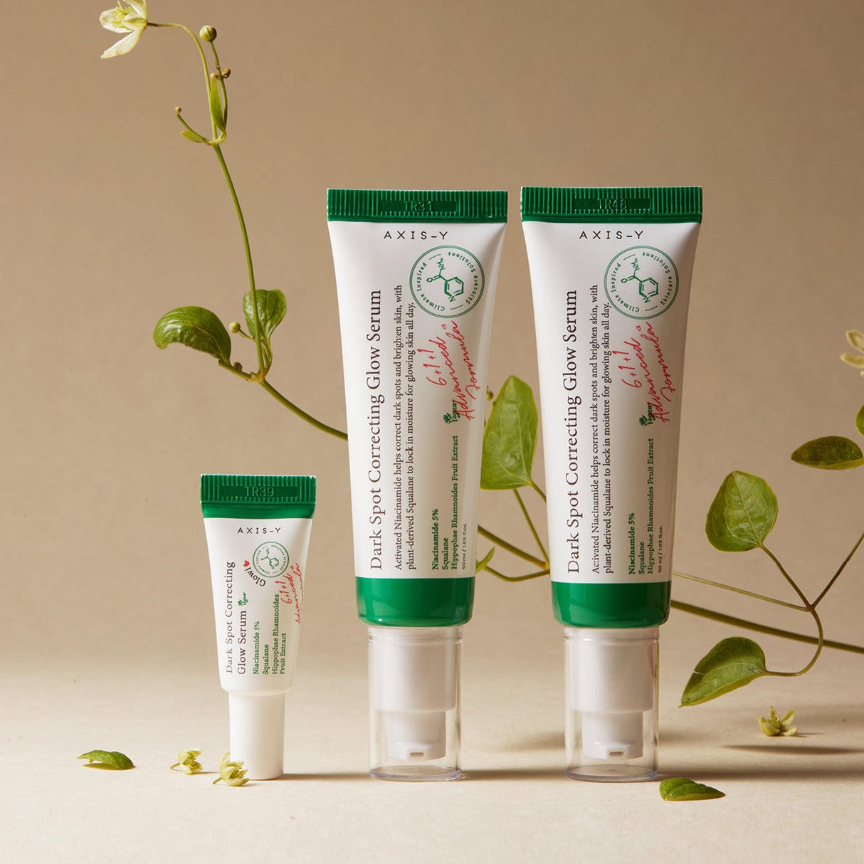 Tre tubetti di siero AXIS-Y Dark Spot Correcting Glow Serum in formati diversi, disposti in verticale su sfondo beige con decorazioni naturali e rampicanti verdi, che trasmettono un’estetica botanica e naturale del brand.

