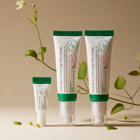 Tre tubetti di siero AXIS-Y Dark Spot Correcting Glow Serum in formati diversi, disposti in verticale su sfondo beige con decorazioni naturali e rampicanti verdi, che trasmettono un’estetica botanica e naturale del brand.

