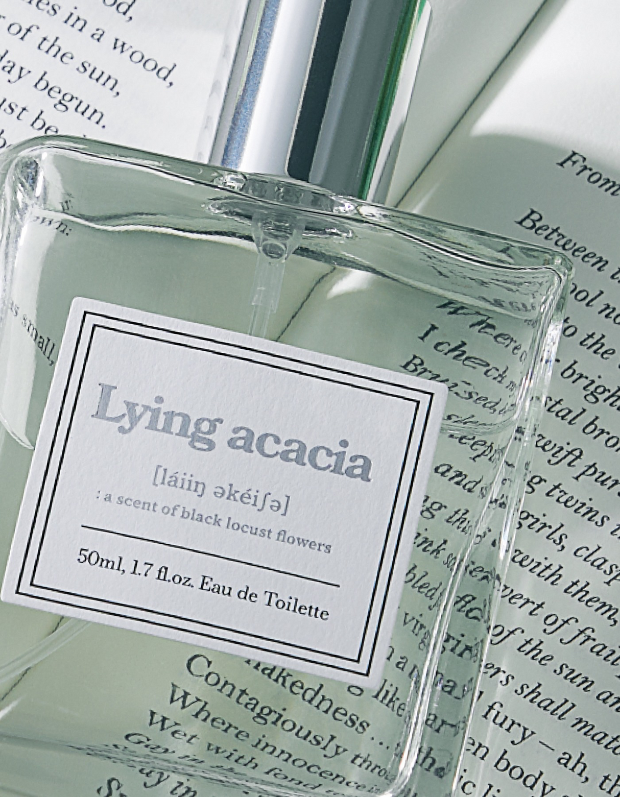 Primo piano del flacone del profumo coreano Ahro Lying Acacia in vetro trasparente con etichetta frontale visibile, fotografato su pagine di libro aperto con luce naturale.