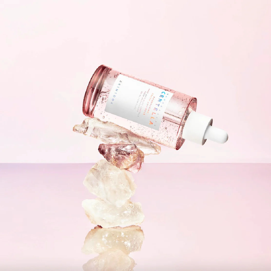 Flacone del siero SKIN1004 Madagascar Centella Pink Ampoule adagiato su una pila di cristalli naturali trasparenti, con sfondo rosa chiaro.