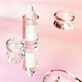 Flacone trasparente con pipetta del siero SKIN1004 Madagascar Centella Pink Ampoule posizionato su superficie rosa riflettente, circondato da gocce decorative in vetro.