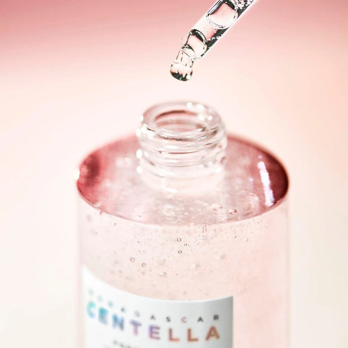 Primo piano della pipetta che rilascia una goccia del siero SKIN1004 Madagascar Centella Pink Ampoule in un flacone aperto, su sfondo rosa tenue.