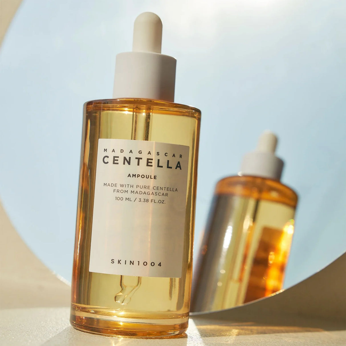 Flacone trasparente da 100ml della ampoule SKIN1004 Madagascar Centella con liquido dorato e tappo bianco, riflesso su superficie lucida