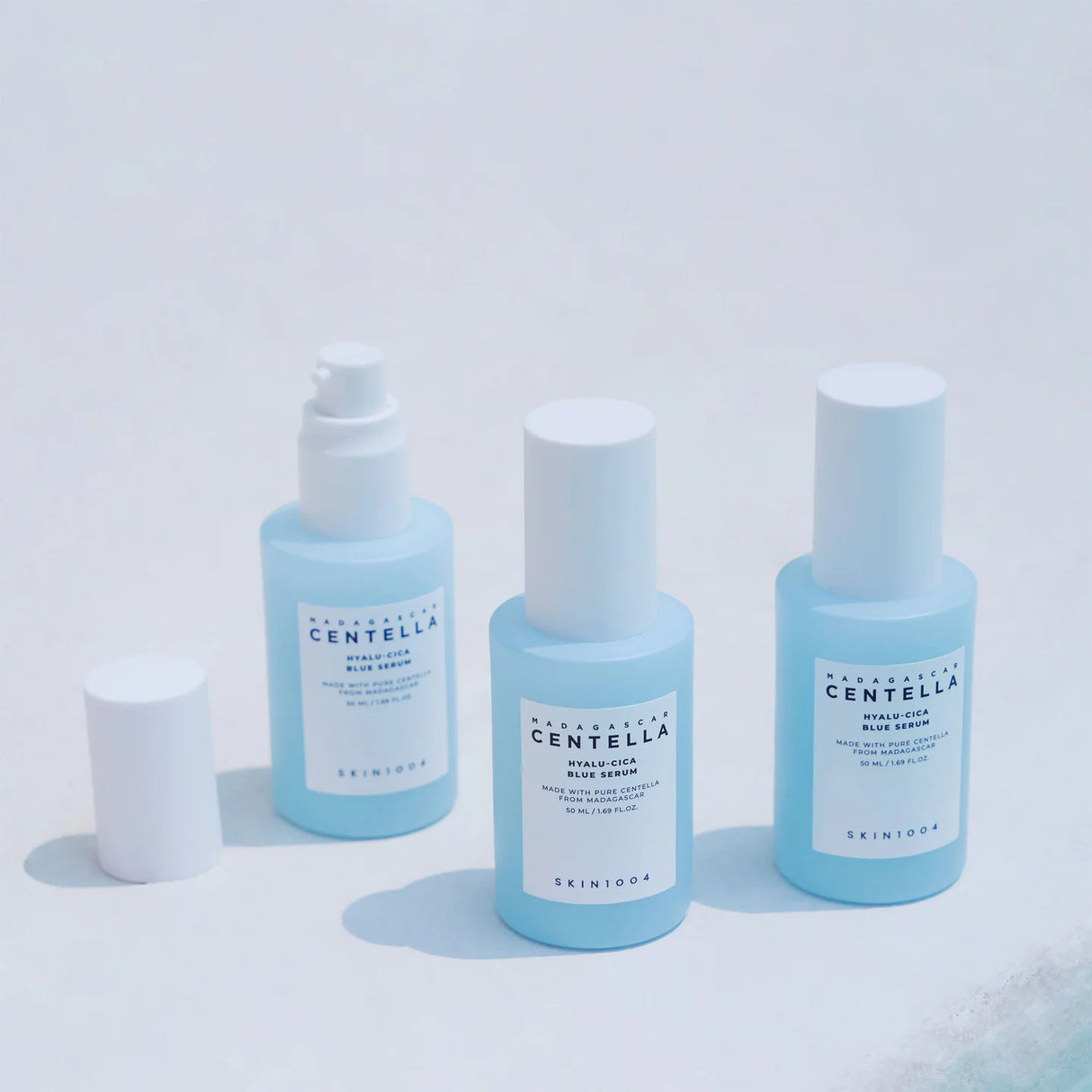 Tre flaconi azzurri della linea SKIN1004 Centella Hyalu-Cica su sfondo bianco, con uno dei flaconi aperto e il tappo appoggiato a lato.
