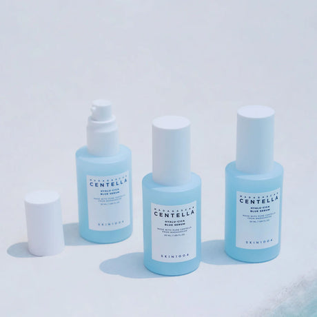 Tre flaconi azzurri della linea SKIN1004 Centella Hyalu-Cica su sfondo bianco, con uno dei flaconi aperto e il tappo appoggiato a lato.