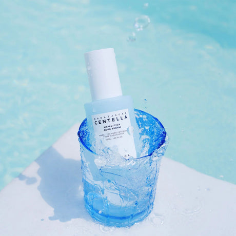 Flacone del siero SKIN1004 Centella Hyalu-Cica Blu Serum immerso in un bicchiere d’acqua trasparente con schizzi, su sfondo azzurro.