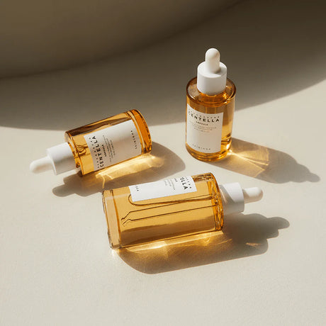 Tre flaconi da 100ml della ampoule SKIN1004 Madagascar Centella con liquido dorato e tappo bianco, disposti su sfondo beige illuminato da luce naturale