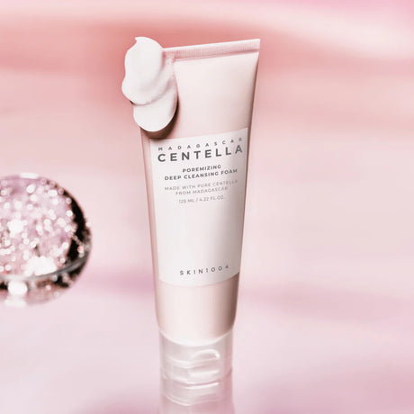 Tubo da 125ml del detergente SKIN1004 Madagascar Centella Poreminizing Deep Cleansing Foam con una dose di prodotto schiumoso sulla parte superiore, su sfondo rosa perlato.