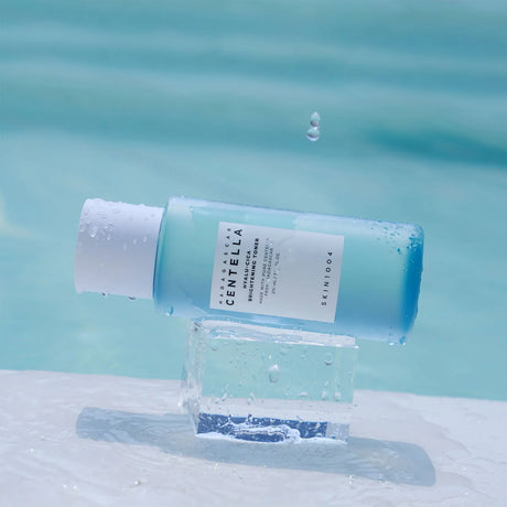 Flacone azzurro da 210ml del tonico SKIN1004 Centella Hyalu-Cica Brightening sdraiato su un cubo di ghiaccio trasparente, con sfondo acquatico azzurro