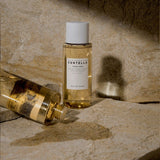 Due flaconi del tonico SKIN1004 Madagascar Centella Toning Toner, uno in piedi e uno sdraiato con tappo svitato e liquido versato, su superficie rocciosa beige con sfondo naturale.
