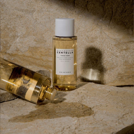 Due flaconi del tonico SKIN1004 Madagascar Centella Toning Toner, uno in piedi e uno sdraiato con tappo svitato e liquido versato, su superficie rocciosa beige con sfondo naturale.

