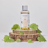 Flacone di Mixsoon Soondy Centella Asiatica Essence posizionato su tre dischi di legno grezzo impilati, circondato da foglie fresche di centella asiatica. Lo sfondo è bianco neutro, che mette in risalto gli elementi naturali del prodotto.

