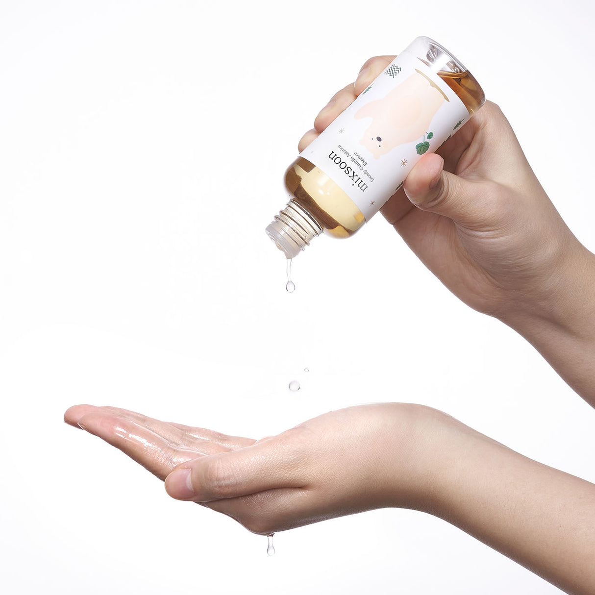 Mano che versa alcune gocce di Mixsoon Soondy Centella Asiatica Essence su un palmo aperto. La bottiglia è inclinata e decorata con un orso stilizzato. Sfondo bianco, atmosfera pulita e minimalista.

