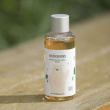 Flacone di Mixsoon Soondy Centella Asiatica Essence posizionato su una superficie in legno all’aperto, con sfondo sfocato verde. Il flacone presenta un’etichetta decorata con un orsetto beige stilizzato e simboli naturali come alberi e foglie.

