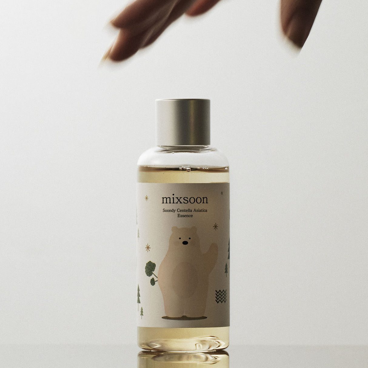 Flacone di Mixsoon Soondy Centella Asiatica Essence in posizione verticale su una superficie riflettente, con due mani sfocate che si avvicinano dall’alto. Il packaging mostra un orsetto stilizzato e dettagli grafici ispirati alla natura.

