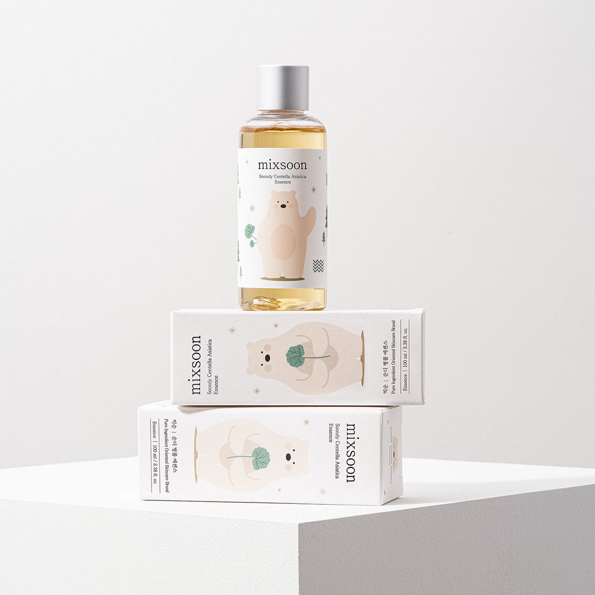 Flacone di Mixsoon Soondy Centella Asiatica Essence posizionato in verticale sopra due scatole coricate dello stesso prodotto. Il packaging presenta un orsetto stilizzato con una foglia verde e grafiche minimaliste su sfondo bianco.

