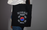 Shopper bag nera in tessuto indossata a spalla da una persona, con grafica Korea Seoul e bandiera sudcoreana centrale su sfondo chiaro