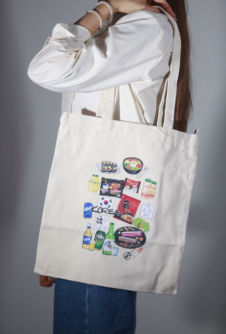 Shopper bag in tessuto color panna portata a spalla da una persona, con grafiche illustrate di ramen, sushi, bevande e snack coreani su sfondo neutro

