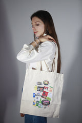 Modella di profilo con capelli lunghi che porta a spalla una shopper bag in tessuto panna con grafiche illustrate di ramen, snack e bevande coreane su sfondo neutro
