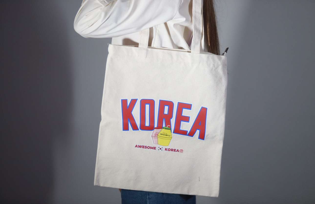 Shopper bag color panna portata a spalla da una persona, con grafica KOREA rossa e blu e piccola illustrazione centrale su sfondo neutro