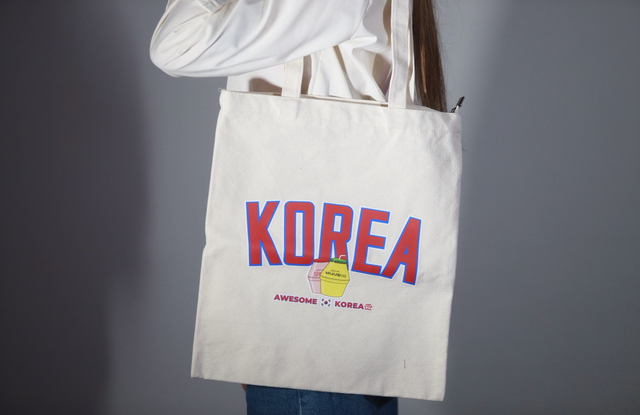 Shopper bag color panna portata a spalla da una persona, con grafica KOREA rossa e blu e piccola illustrazione centrale su sfondo neutro