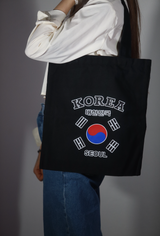 Tote bag nera in tessuto portata a spalla da una persona con camicia bianca, con grafica Korea Seoul e bandiera della Corea del Sud stampata al centro
