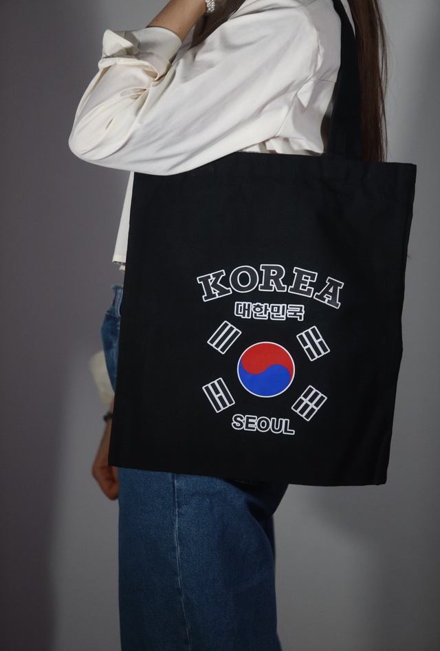 Tote bag nera in tessuto portata a spalla da una persona con camicia bianca, con grafica Korea Seoul e bandiera della Corea del Sud stampata al centro
