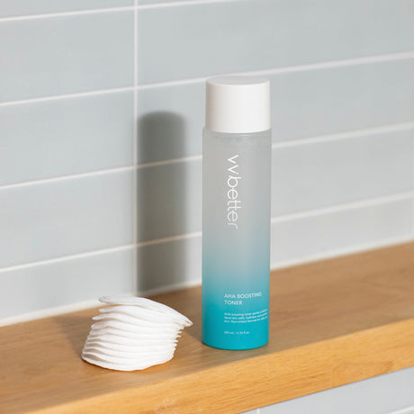 Flacone verticale sfumato azzurro del tonico "AHA Boosting Toner" di vvbetter da 200 ml, posizionato su una mensola in legno chiaro accanto a una pila ordinata di dischetti di cotone, con sfondo piastrellato chiaro.