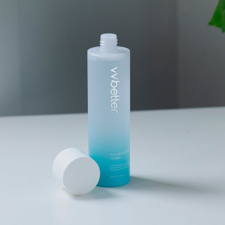 Flacone sfumato azzurro del tonico "AHA Boosting Toner" di vvbetter da 200 ml, con tappo bianco rimosso appoggiato accanto, su sfondo minimal bianco e grigio.