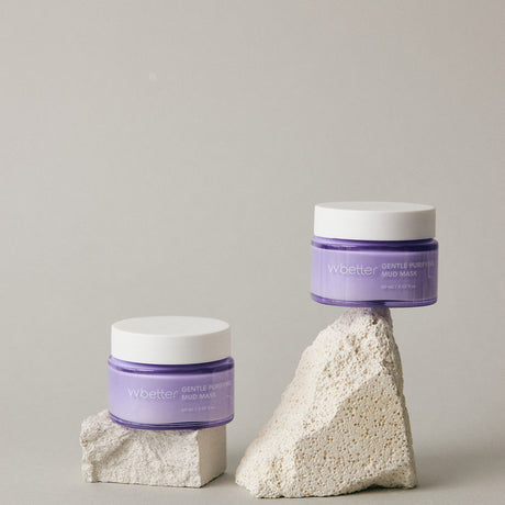 Due vasetti del vvbetter Gentle Purifying Mud Mask in vetro lilla con coperchio bianco, elegantemente posizionati su blocchi di pietra beige. Lo sfondo neutro e minimalista enfatizza il packaging del prodotto, trasmettendo un senso di purezza e cura della pelle.