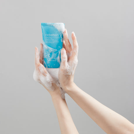 Due mani insaponate tengono il tubo azzurro di vvbetter 5.5 Soothing Cleansing Foam contro uno sfondo grigio chiaro, mettendo in risalto la texture schiumosa del prodotto e la sua funzione detergente delicata.