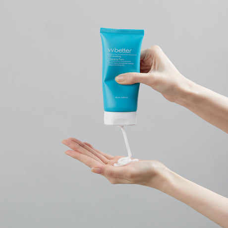 Una mano tiene il tubo azzurro del vvbetter 5.5 Soothing Cleansing Foam mentre l’altra riceve una dose del detergente bianco cremoso, mostrando la consistenza del prodotto contro uno sfondo grigio chiaro.