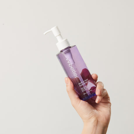 Mano che tiene un flacone trasparente color lavanda del detergente viso vvbetter Gentle Deep Cleansing Oil con dispenser bianco, su sfondo neutro.