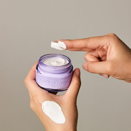 Mani che applicano la vvbetter Gentle Purifying Mud Mask da un barattolo lilla. Una mano regge il contenitore aperto, mentre l’altra raccoglie una generosa quantità di maschera con l’indice. Sulla parte superiore della mano che regge il barattolo è già stesa una porzione del prodotto, mostrando la consistenza cremosa e densa.